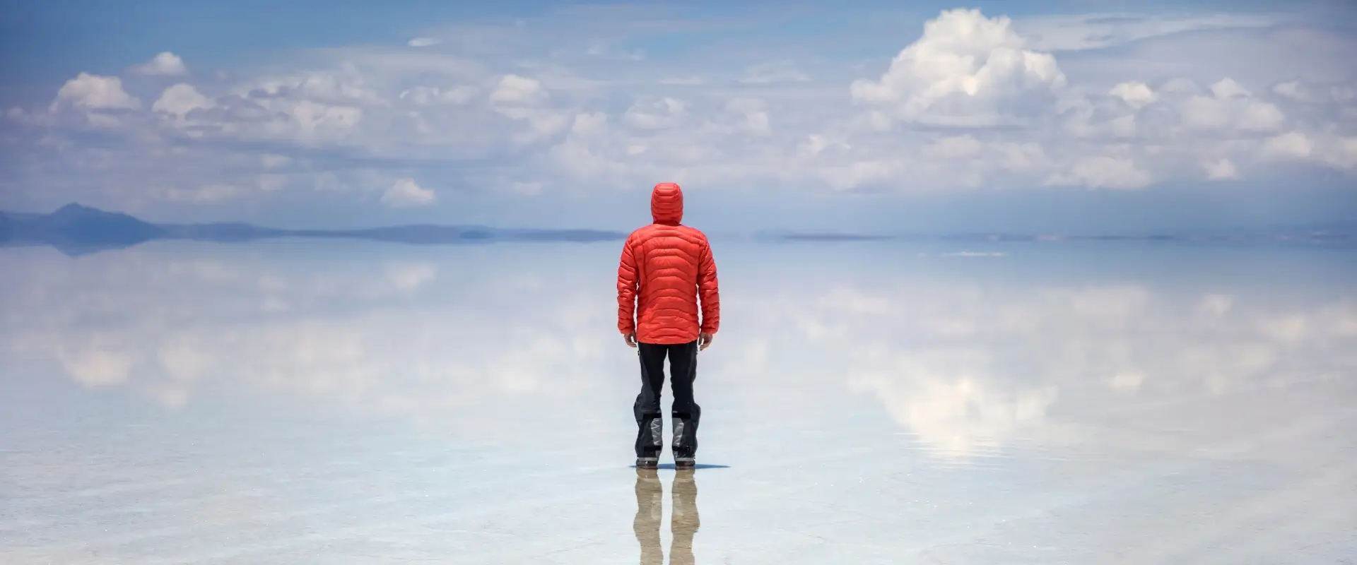 Um homem de costas, de jaqueta vermelha e calças pretas no meio de um lagoa razo e tranquilo, refletindo o céu azul claro como se fosse um espelho, observando o horizonte desse lago com algumas montanhas em Salar de Uyuni.